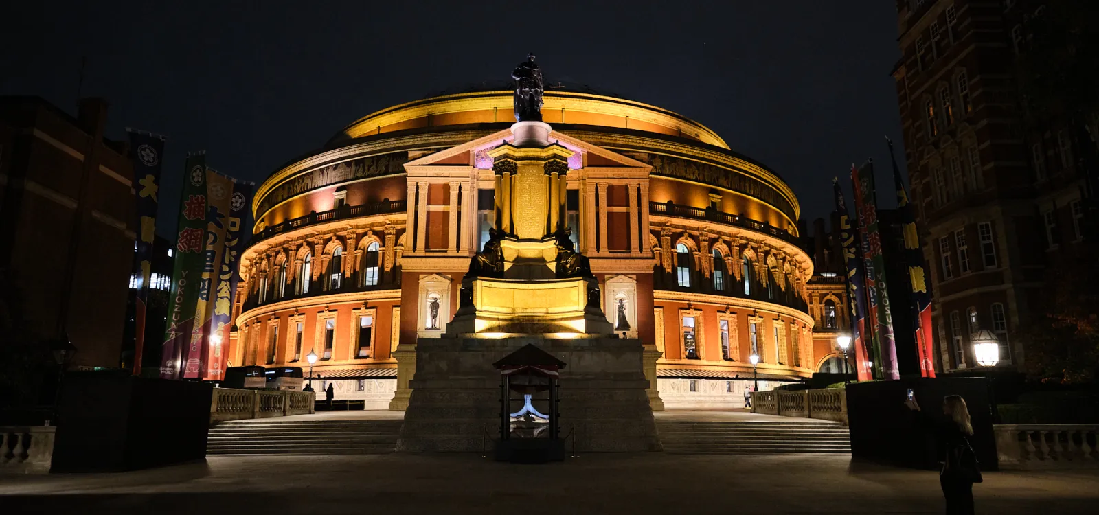 Royal Albert Hall illuminated at night, with the Albert Memorial in the foreground and golden architectural lighting against the dark London sky.