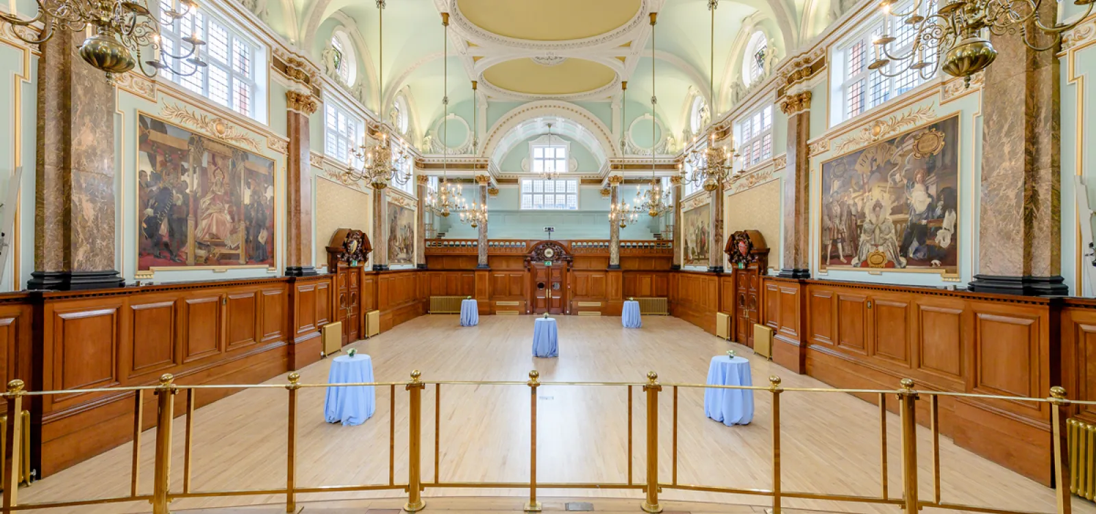 The Main Hall at Chelsea Old Town Hall, featuring ornate gold detailing, high arched ceilings, large windows, wood panelling, historic murals and elegant chandeliers.