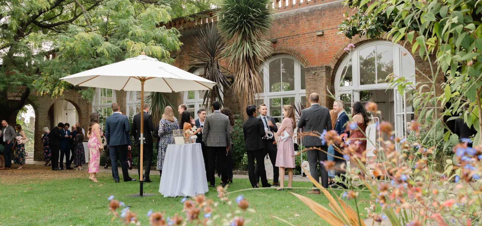 Guests on the lawn at The Orangery, Holland Park, surrounded by lush greenery, mature trees and the venue’s historic brick arches.