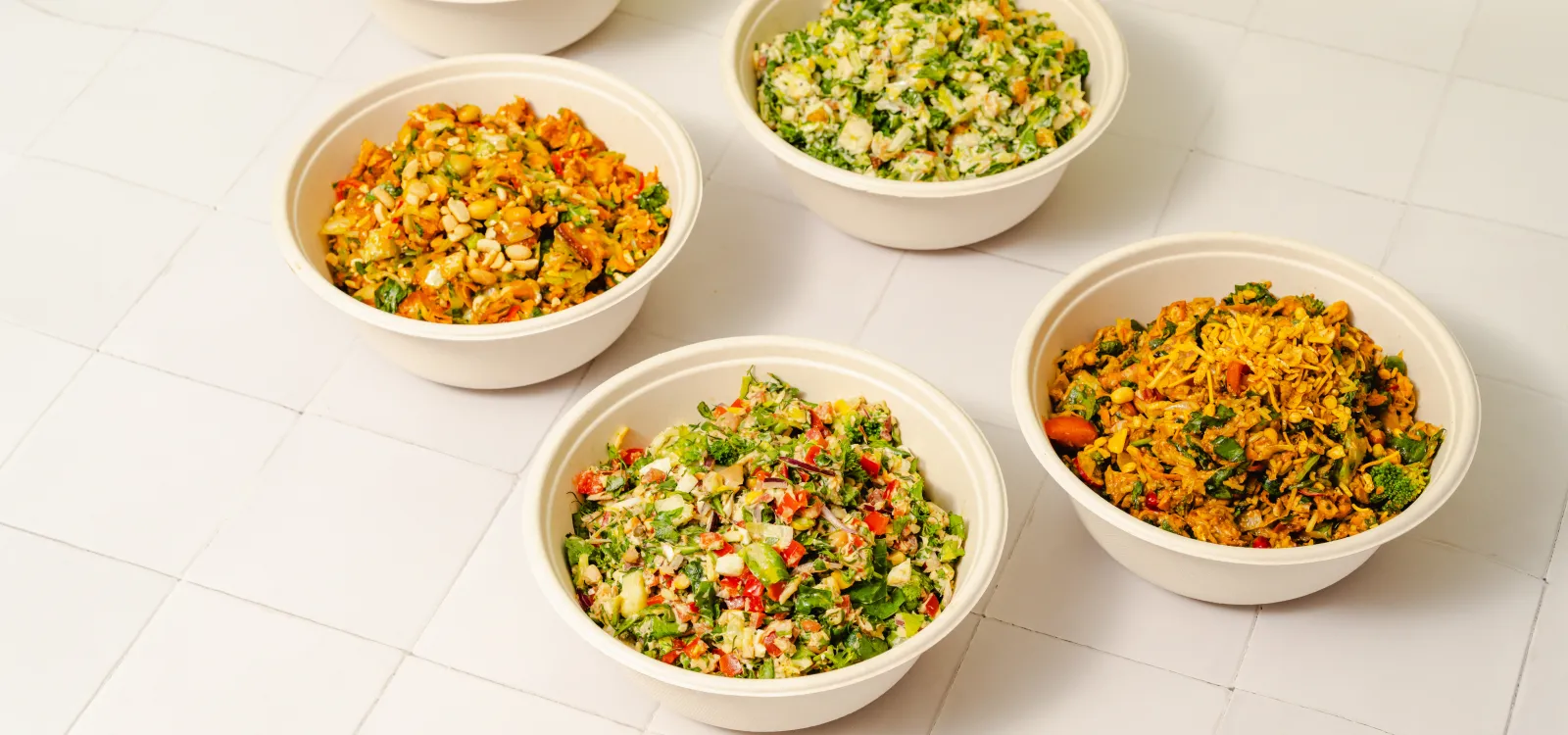 Overhead view of five freshly chopped salad bowls featuring colourful vegetables, greens and mixed toppings on a cream tiled surface.