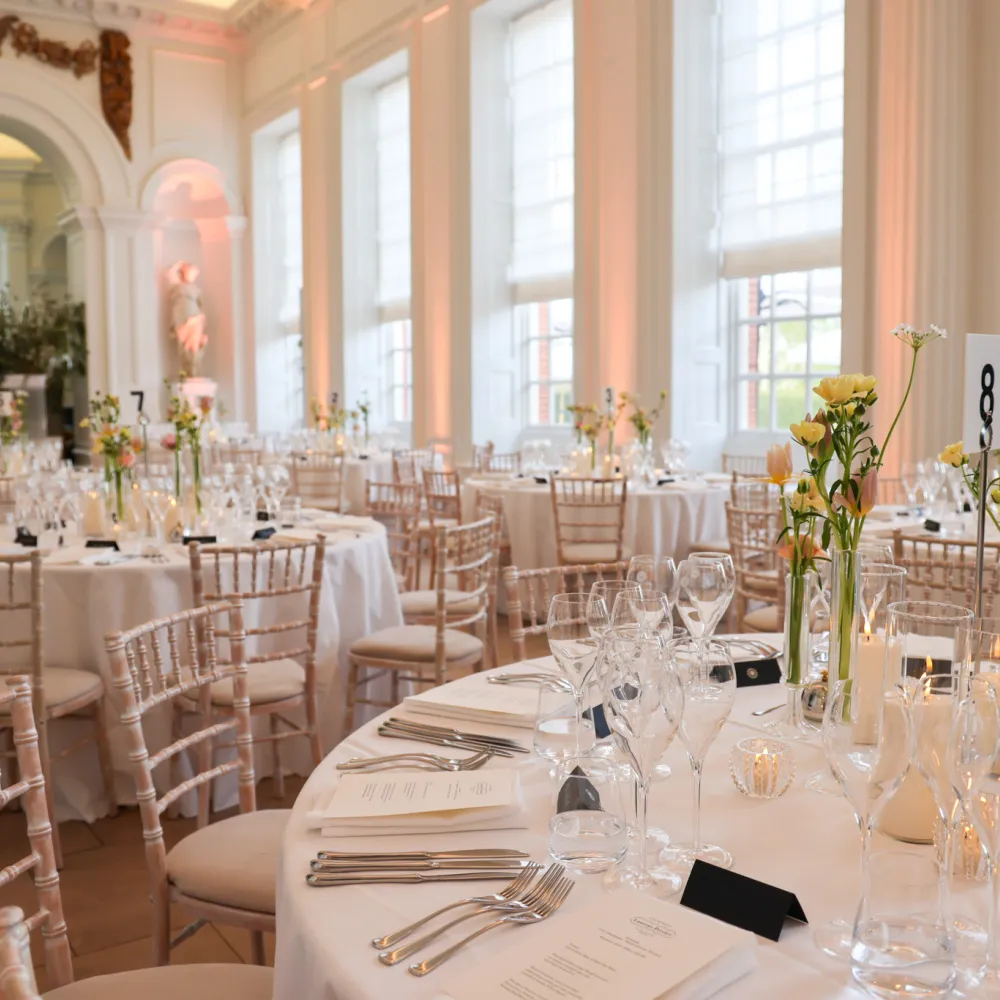 Round tables set with white linens, glassware, candles, and floral centrepieces in the Orangery Room at Kensington Palace
