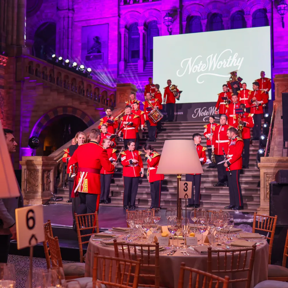 Dinner set up in Hintze Hall at the Natural History Museum, round tables, the band in red uniforms on the staircase, and the suspended blue whale skeleton above.