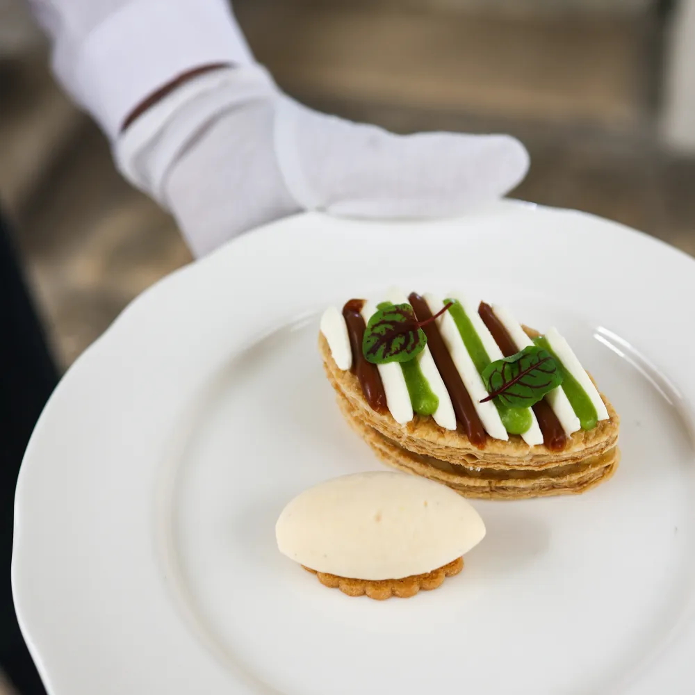 A server presents Rhubarb's Apple & Miso Mille Feuille dessert, featuring caramelised Granny Smith apple, miso caramel, and Tahitian vanilla ice cream at an anniversary lunch at Queen's House, Greenwich.