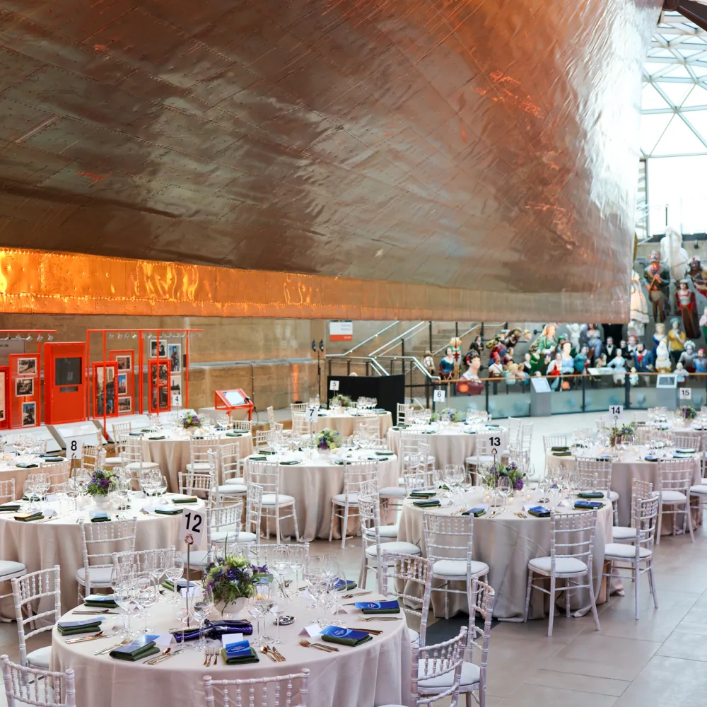 **Image Name:** Dinner Beneath the Cutty Sark  **Alt Text:** Elegantly dressed dining tables arranged beneath the Cutty Sark's iconic copper hull in the Dry Berth, creating a spectacular setting for a private dinner in Greenwich.