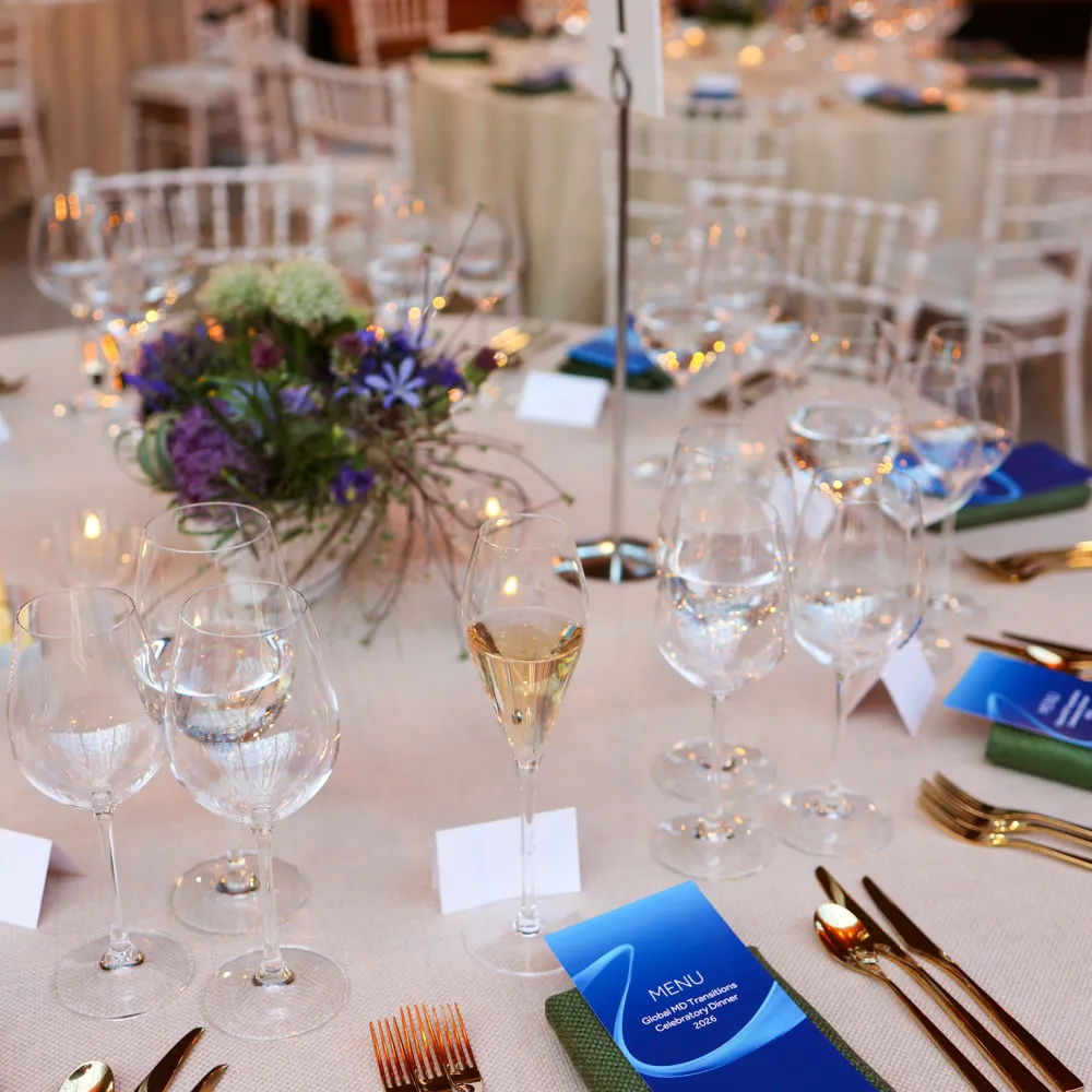 Elegantly dressed dining tables beneath the Cutty Sark, featuring floral centrepieces in blue and purple tones, gold cutlery, branded menus, and white chairs arranged for a private dinner reception.