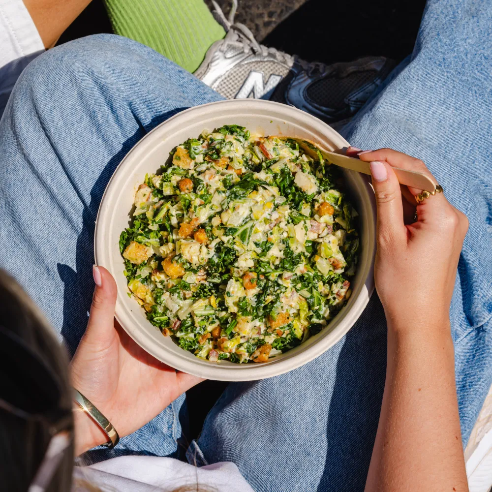 Customer holding a bowl of creamy chopped chicken Caesar salad while seated outdoors in the sunshine.