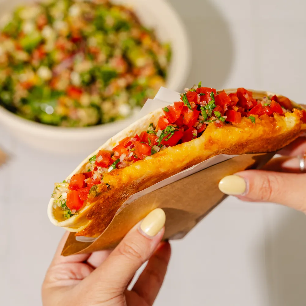 : Close-up of a toasted wrap filled with golden melted cheese and fresh chopped tomatoes, held in hand with a salad bowl in the background.