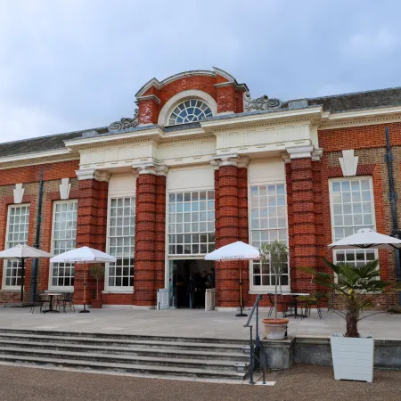 Exterior of the Orangery at Kensington Palace, a red-brick historic building with tall windows and outdoor seating with branded parasols