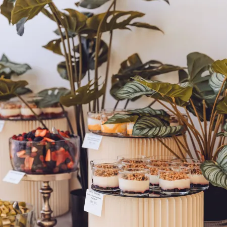 Breakfast catering display featuring yogurt parfaits with granola, fresh fruit bowls, and decorative greenery arranged on tiered stands