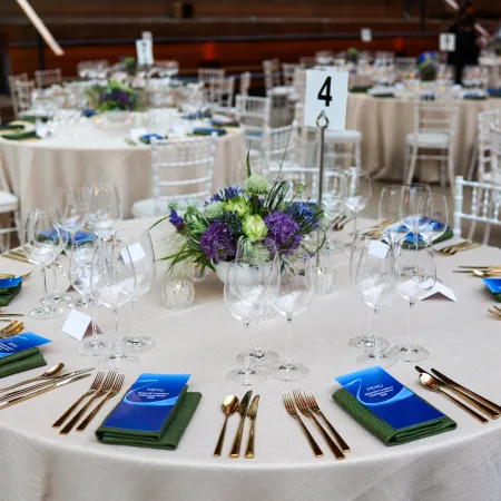 Elegantly dressed dining tables beneath the Cutty Sark, featuring floral centrepieces in blue and purple tones, gold cutlery, branded menus, and white chairs arranged for a private dinner reception.