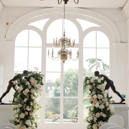 The Orangery at Holland Park featuring dramatic arched windows, ornate chandeliers and floral arrangements framing the ceremony aisle, with white seating and views of the surrounding gardens.