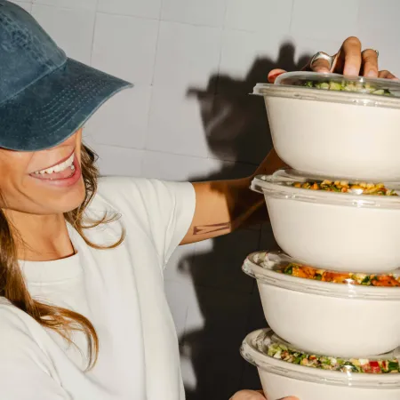 Smiling customer in a denim cap holding a stack of takeaway salad bowls with fresh ingredients.