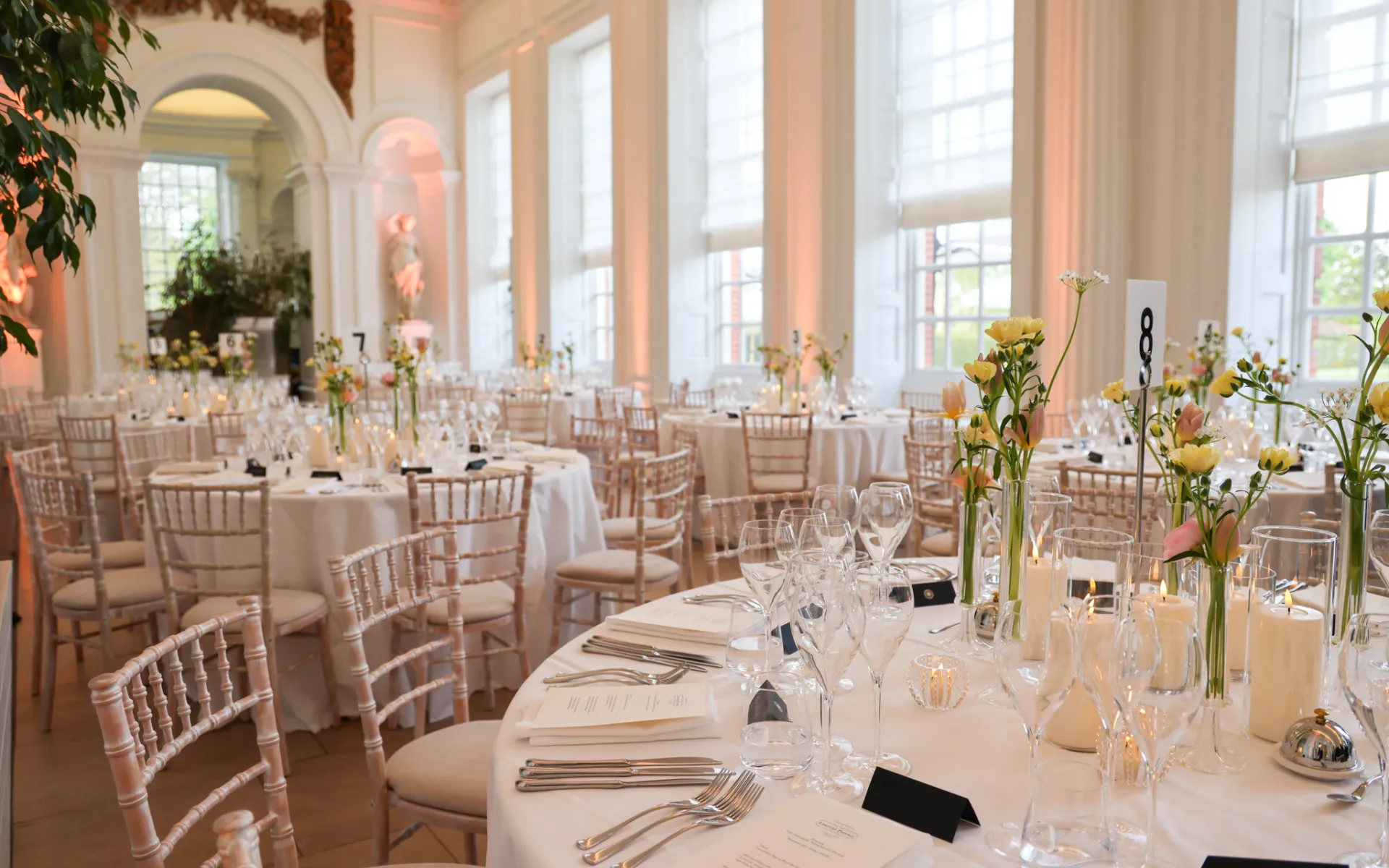 Round tables set with white linens, glassware, candles, and floral centrepieces in the Orangery Room at Kensington Palace