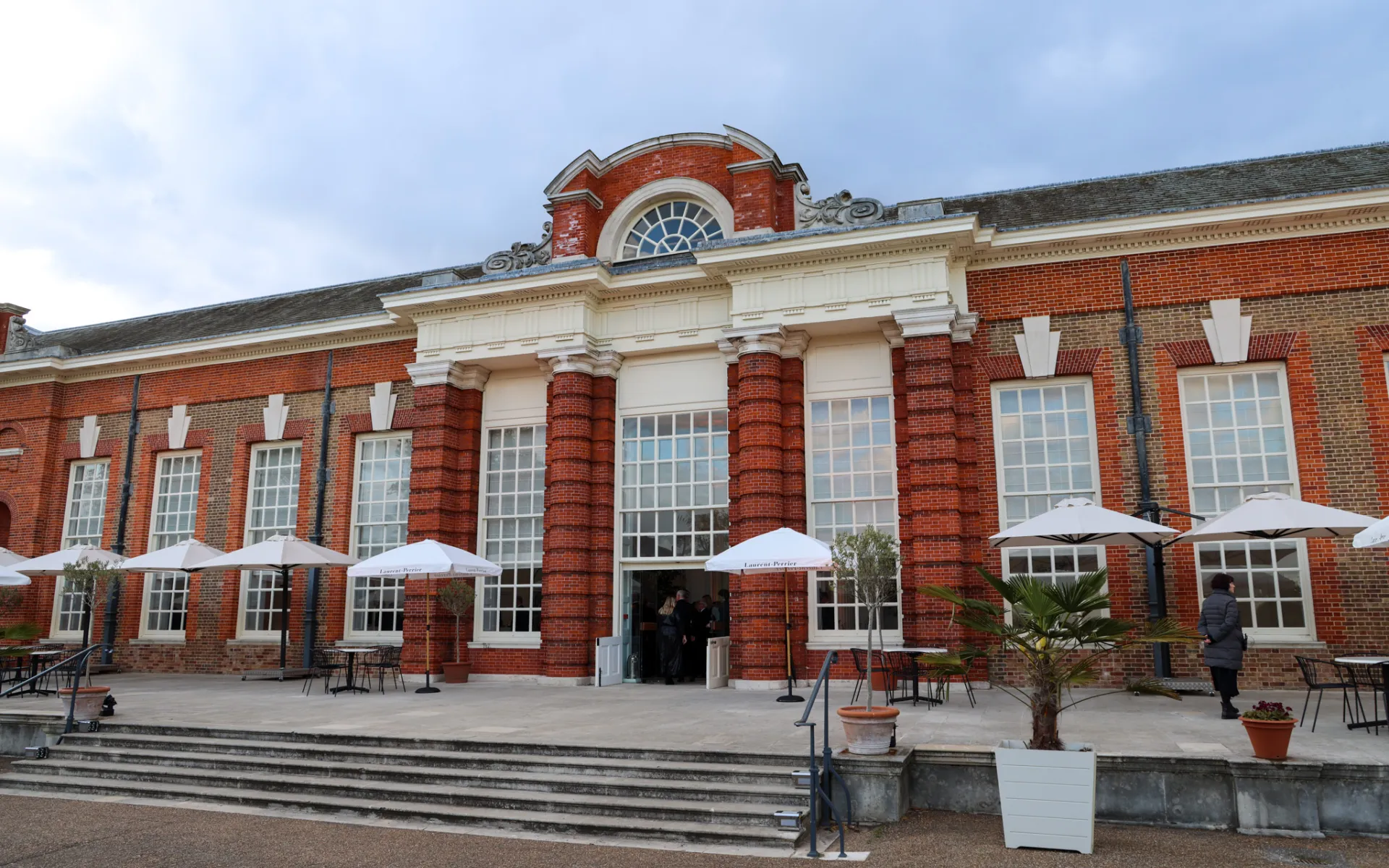 Exterior of the Orangery at Kensington Palace, a red-brick historic building with tall windows and outdoor seating with branded parasols