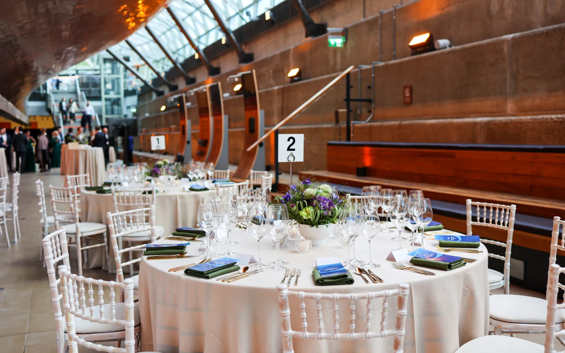 Elegantly dressed dining tables beneath the Cutty Sark, featuring floral centrepieces in blue and purple tones, gold cutlery, branded menus, and white chairs arranged for a private dinner reception.