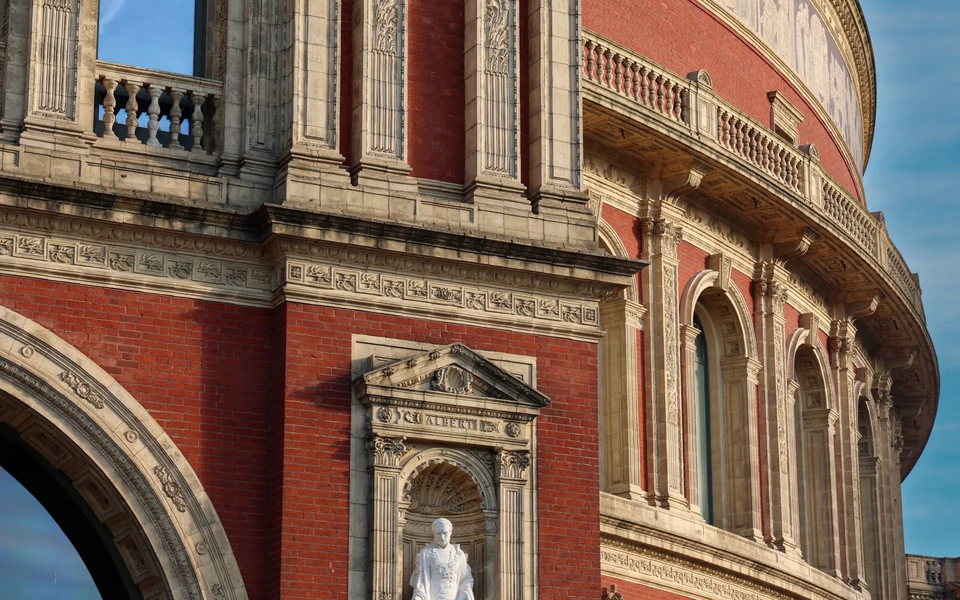 Close-up of the Royal Albert Hall’s ornate red-brick and stone façade, showcasing its intricate architectural details, decorative columns and sculpted niches against a clear blue sky.