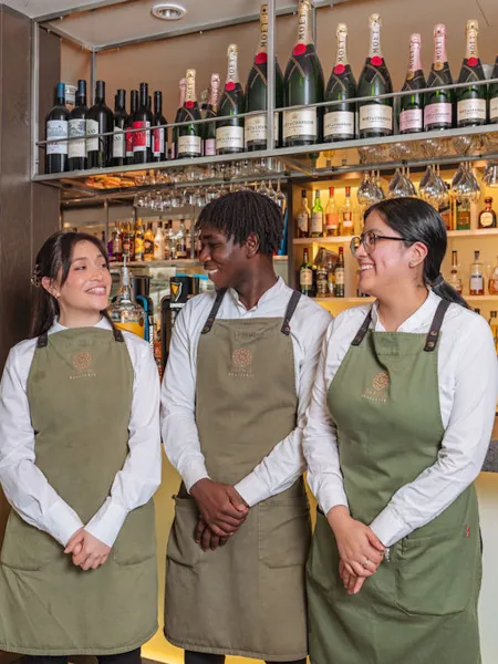 Three members of Rhubarb hospitality bar team standing together behind a bar, smiling and wearing aprons
