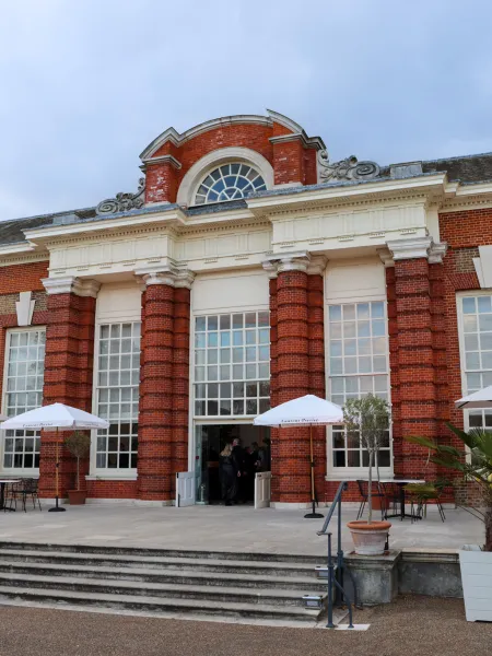 Exterior of the Orangery at Kensington Palace, a red-brick historic building with tall windows and outdoor seating with branded parasols