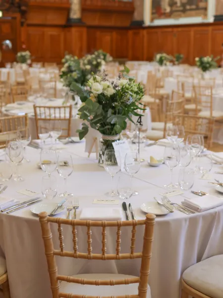 A beautifully dressed dining room at Chelsea Old Town Hall, with round tables set with white linen, glassware and floral centrepieces, surrounded by elegant wood panelling and Chiavari chairs.
