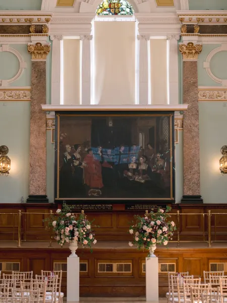 The Main Hall at Chelsea Old Town Hall, featuring ornate chandeliers, marble columns, gilded detailing and a historic painting, with floral arrangements and white chairs set up for a ceremony.