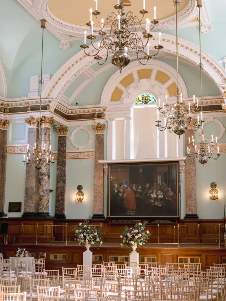 The Main Hall at Chelsea Old Town Hall, featuring ornate chandeliers, marble columns, gilded detailing and a historic painting, with floral arrangements and white chairs set up for a ceremony.
