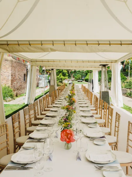 Long dining tables set beneath a marquee at The Orangery, Holland Park, with natural wood chairs, colourful floral centrepieces and garden views.