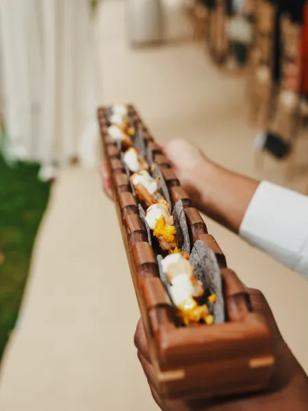 A close-up of a canapé served in a wooden presentation tray, featuring dark crisp shells, creamy toppings and vibrant orange garnish.