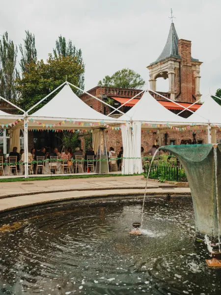 Outdoor event setup at The Orangery, Holland Park, with guests dining beneath white marquees beside the garden fountain and historic brick architecture.