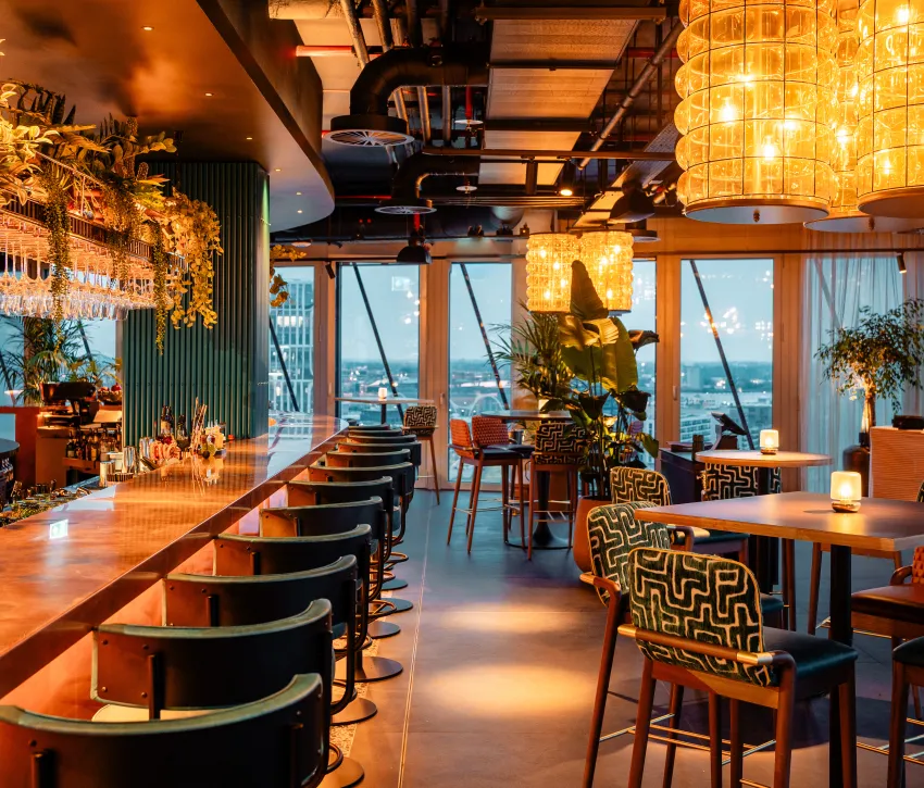 Warmly lit bar interior with high tables, lounge seating, tables and greenery at AirBar13.