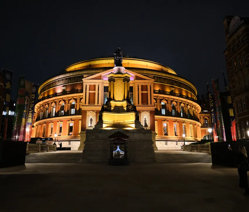 Royal Albert Hall illuminated at night, with the Albert Memorial in the foreground and golden architectural lighting against the dark London sky.