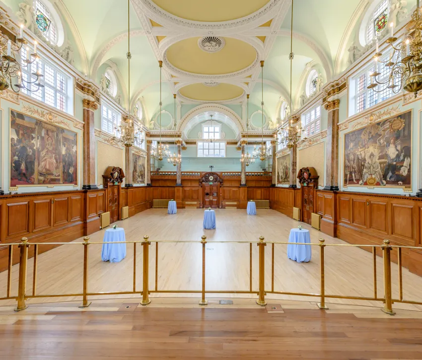 The Main Hall at Chelsea Old Town Hall, featuring ornate gold detailing, high arched ceilings, large windows, wood panelling, historic murals and elegant chandeliers.