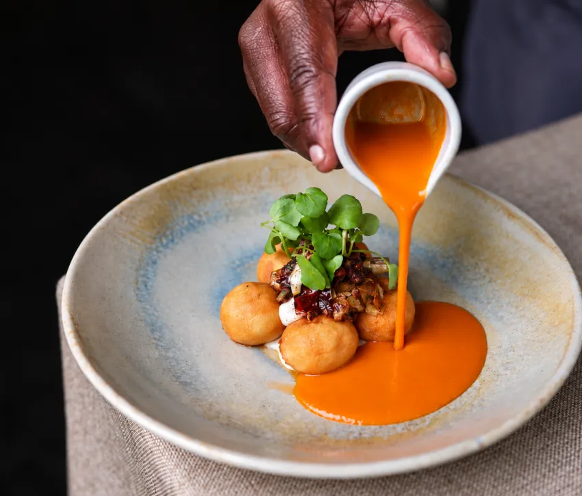Festive gnocchi served with a rich orange sauce, topped with microgreens and garnished with a creamy sauce and finely chopped ingredients, captured as the sauce is poured over the dish.