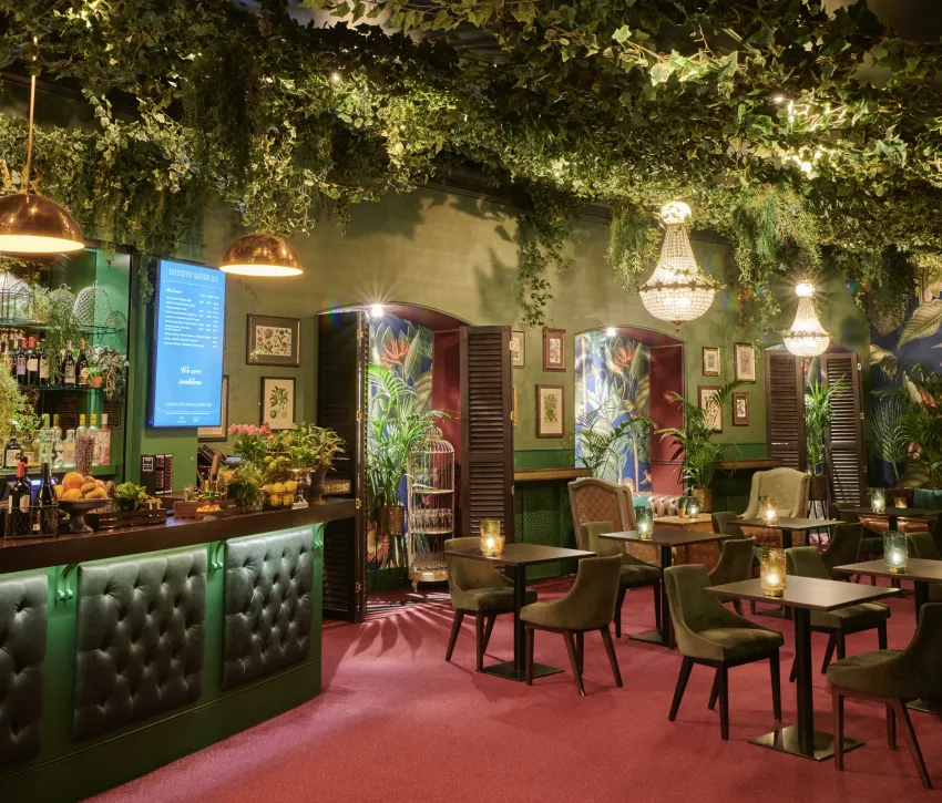 Interior of a vibrant Kensington Gardens Bar dining space, featuring lush overhead greenery, emerald-green bar seating, statement chandeliers, tropical artwork and warm ambient lighting.