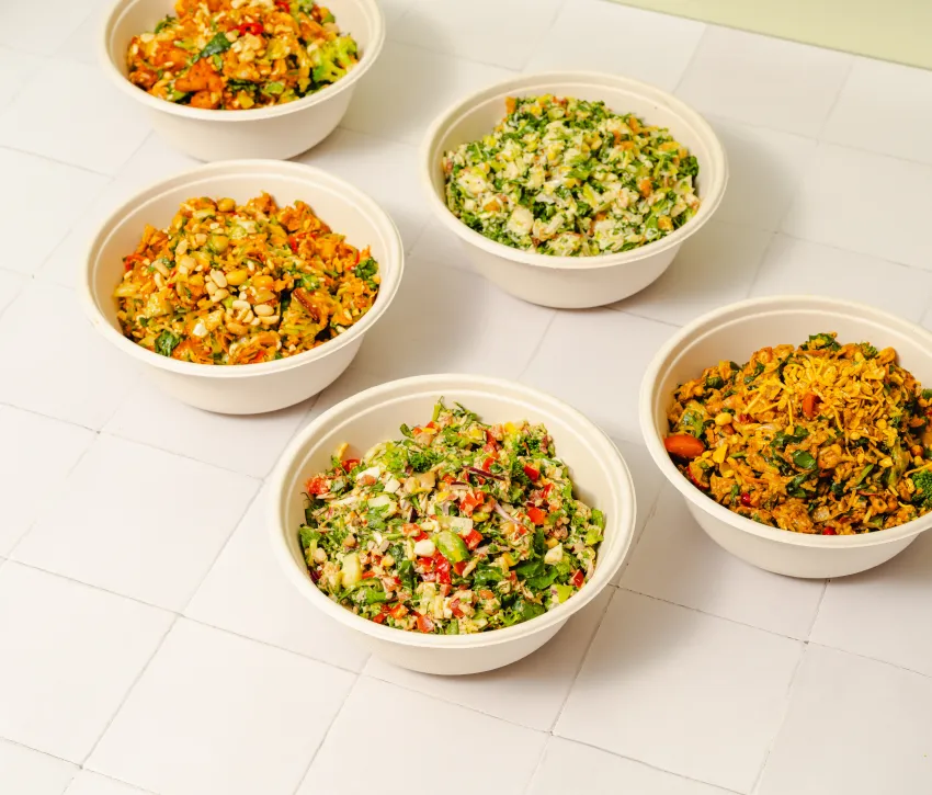 Overhead view of five freshly chopped salad bowls featuring colourful vegetables, greens and mixed toppings on a cream tiled surface.