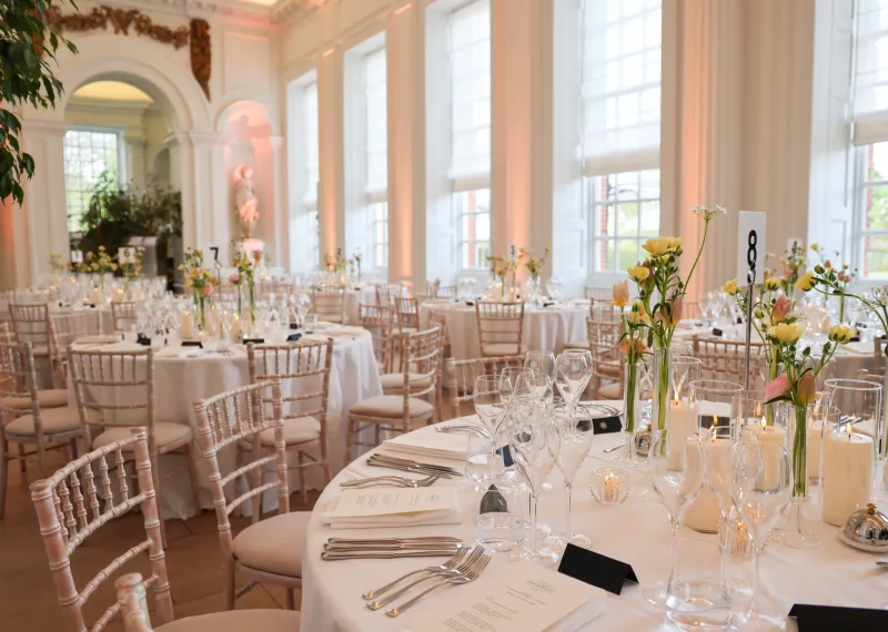 Round tables set with white linens, glassware, candles, and floral centrepieces in the Orangery Room at Kensington Palace
