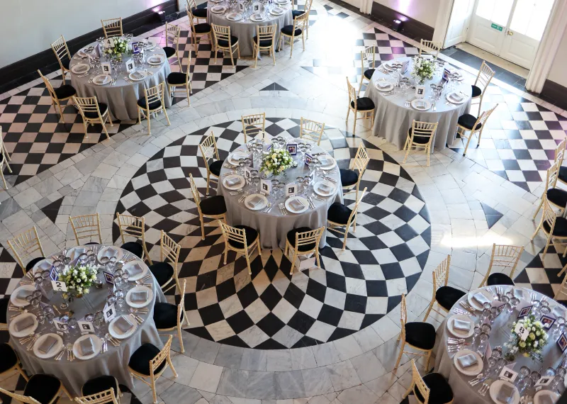 Round dining tables elegantly arranged in the Great Hall at Queen's House, Greenwich, featuring floral centrepieces, silver table settings, and gold chairs against the venue's iconic black-and-white geometric marble floor.