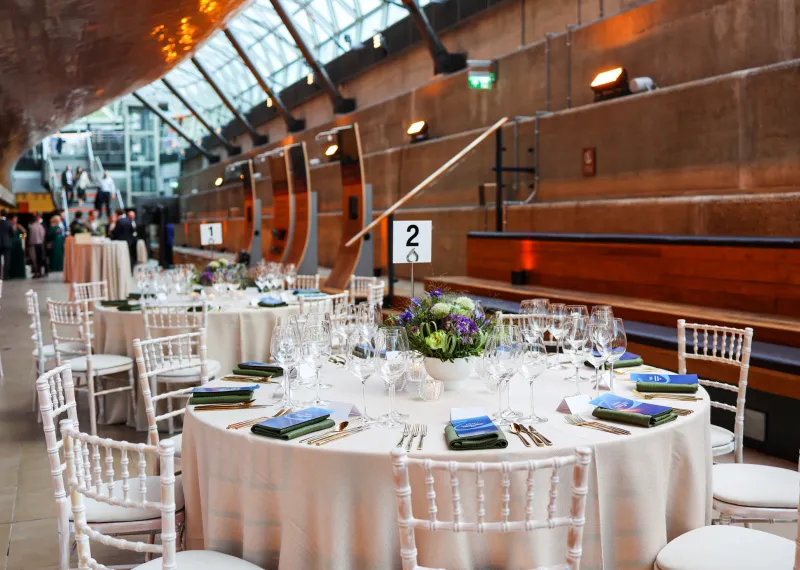 Elegantly dressed dining tables beneath the Cutty Sark, featuring floral centrepieces in blue and purple tones, gold cutlery, branded menus, and white chairs arranged for a private dinner reception.