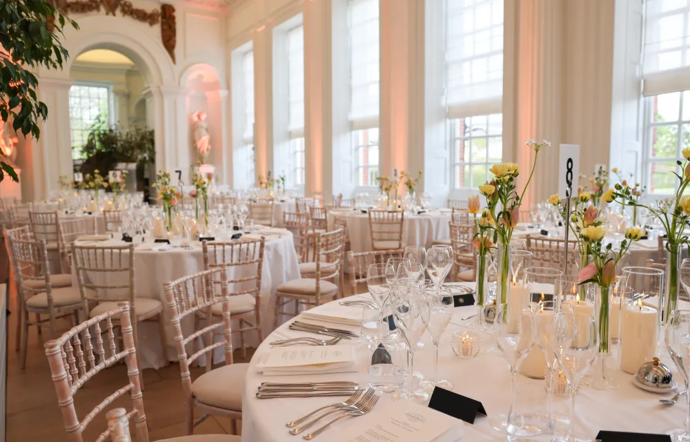 Round tables set with white linens, glassware, candles, and floral centrepieces in the Orangery Room at Kensington Palace