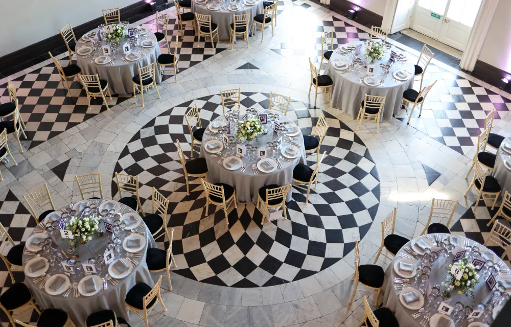 Round dining tables elegantly arranged in the Great Hall at Queen's House, Greenwich, featuring floral centrepieces, silver table settings, and gold chairs against the venue's iconic black-and-white geometric marble floor.