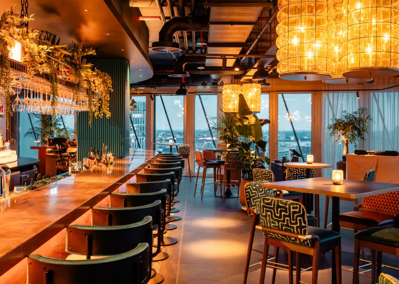 Warmly lit bar interior with high tables, lounge seating, tables and greenery at AirBar13.