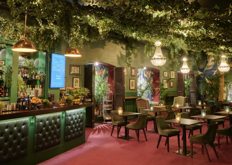 Interior of a vibrant Kensington Gardens Bar dining space, featuring lush overhead greenery, emerald-green bar seating, statement chandeliers, tropical artwork and warm ambient lighting.