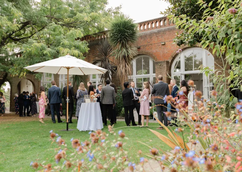 Guests on the lawn at The Orangery, Holland Park, surrounded by lush greenery, mature trees and the venue’s historic brick arches.