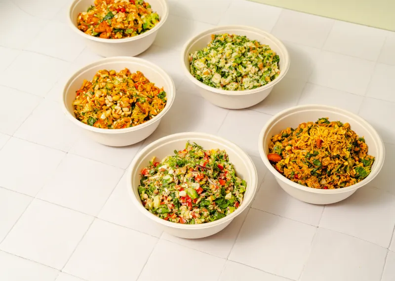 Overhead view of five freshly chopped salad bowls featuring colourful vegetables, greens and mixed toppings on a cream tiled surface.
